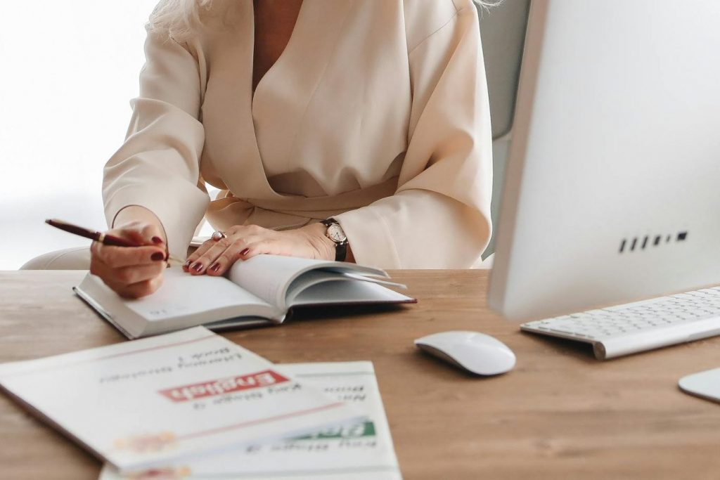 Elegant senior woman writing at office desk with computer, showcasing a professional work environment.