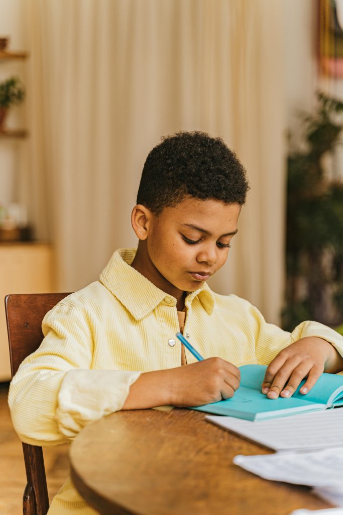 A young boy in a yellow shirt sits at a table taking notes indoors, focused and engaged.